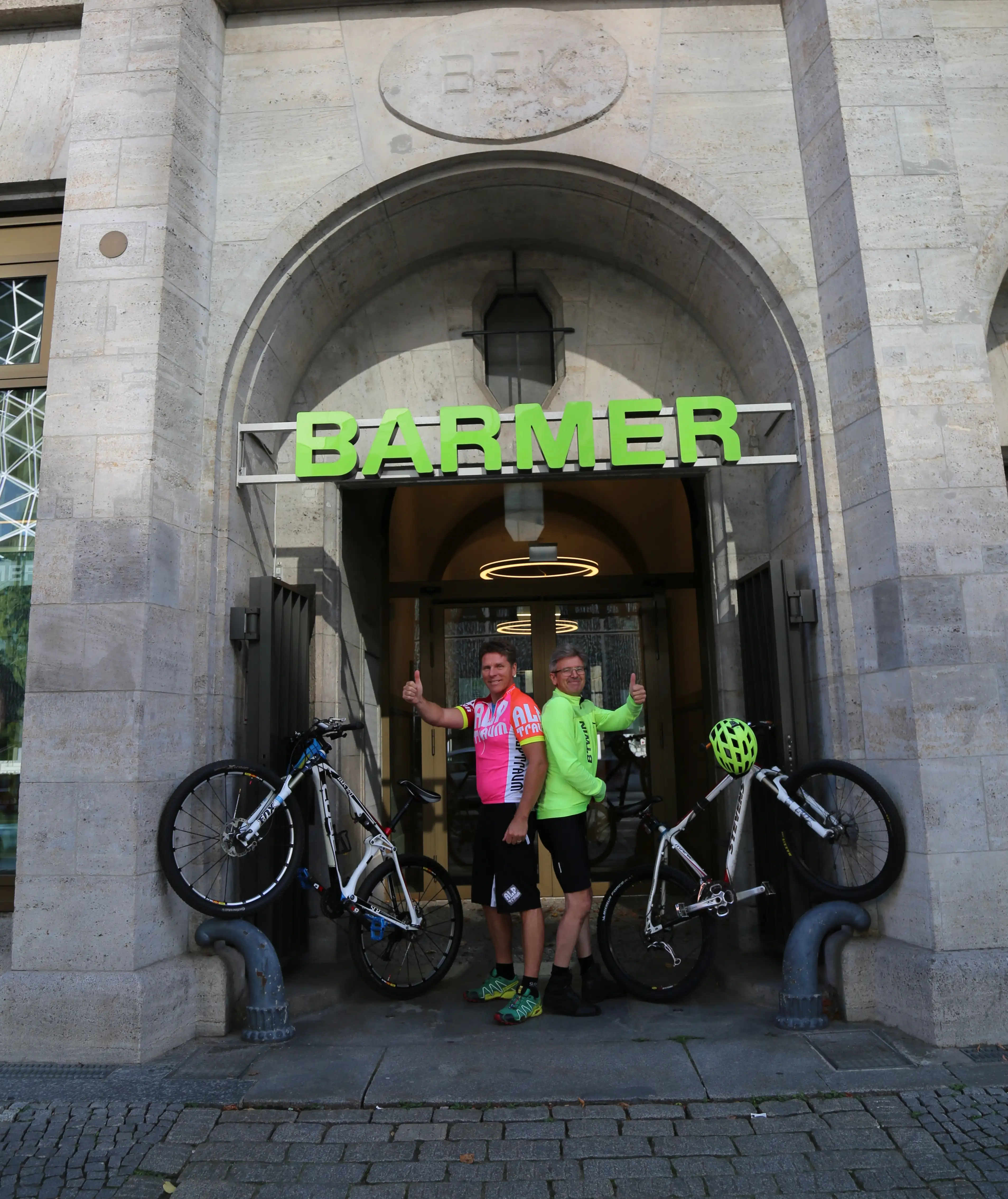 Zwei Barmer Mitarbeiter, die mit ihren Fahrräder ein V in der Eingangspforte von Barmer bilden