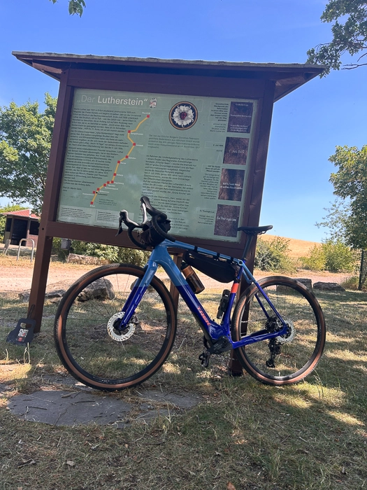 Ein blaues Fahrrad steht vor einer Informationstafel im Schatten eines Baumes. Im Hintergrund sind Bäume und ein klarer blauer Himmel zu sehen.